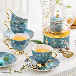 May include: A tea set with a teapot, teacups, saucers, and a spoon. The set has a teal background with a white floral design and gold accents. The teacups are filled with tea. The teapot is made of glass and teal ceramic.