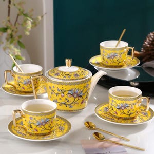 Puede incluir: Un juego de té amarillo con diseños florales. El juego incluye una tetera, cuatro tazas, platillos y cucharas. Las tazas y la tetera tienen asas y detalles dorados. El juego de té está sobre una superficie blanca.