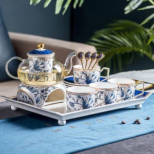Puede incluir: Juego de té de porcelana con tetera de vidrio, cuatro tazas, platillos y cucharas, decorado con diseños florales azules y detalles dorados. El juego incluye una bandeja blanca con borde azul, para servir el té.