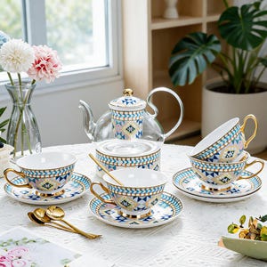 Puede incluir: Una tetera de cristal con infusor y tapa de cerámica a juego, acompañada de tres tazas y platillos de cerámica. El conjunto presenta un patrón geométrico en azul, rosa y dorado. Tres cucharas de tono dorado están sobre la mesa.