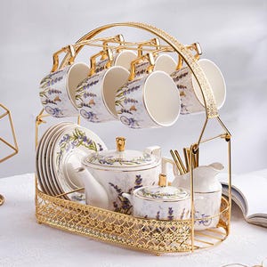 Könnte beinhalten: Goldfarbenes Metall-Teeservice mit passendem Ständer. Das Set umfasst Tassen, Untertassen, eine Teekanne, eine Zuckerdose und einen Sahnekännchen. Die weißen Porzellanstücke sind mit Lavendelzweigen und Fahrradmotiven verziert.