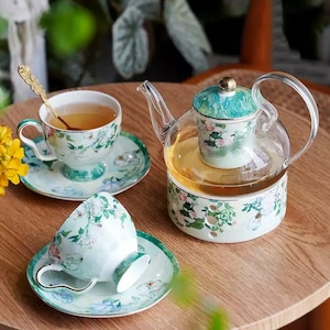 Peut inclure: Service à thé fleuri comprenant une théière en verre et deux tasses avec soucoupes. La théière a un couvercle décoratif, et les tasses et la base de la théière ont un fond vert clair avec des motifs floraux. Une cuillère dorée repose dans une tasse.