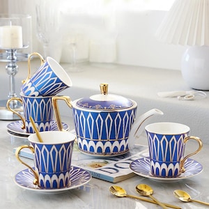 Puede incluir: Un juego de té de porcelana azul y blanco con detalles dorados. El juego incluye una tetera, tazas, platillos y cucharas. Las tazas y la tetera presentan un diseño geométrico. Las asas y los bordes son dorados.