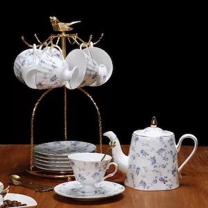 Può includere: Servizio da tè in porcellana bianca con motivi floreali blu. Il set include una teiera, tazze, piattini e un supporto in metallo dorato. Il supporto ha un ornamento a forma di uccello. Il servizio da tè è disposto su una superficie di legno.