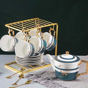 Könnte beinhalten: Ein Teeservice mit einem goldfarbenen Metallgestell, das sechs passende Tassen mit Untertassen hält. Die Tassen und die Teekanne sind weiß mit teal- und goldfarbenen Akzenten. Goldene Löffel und eine Teekanne vervollständigen das Set.