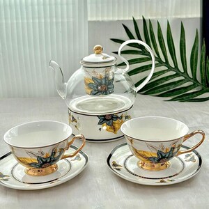 Könnte beinhalten: Elegantes Teeservice mit einer Glasteekanne mit floralem Aufsatz, zwei Tassen und Untertassen. Die weißen Porzellantassen und Untertassen haben goldene Ränder und florale Designs. Die Teekanne steht auf einem weißen Sockel mit passenden floralen Akzenten.