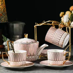 May include: A pink and gold tea set comprising a teapot, sugar bowl, two teacups, saucers, and a gold-coloured metal stand. The teacups and teapot feature a geometric design. The set includes two gold spoons.
