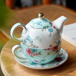 May include: A floral tea set comprising a teapot, teacup, and saucer. The set has a pale blue base with pink and blue floral patterns. The teapot features a gold knob on the lid and gold trim on the handle. The teacup is placed on the saucer.