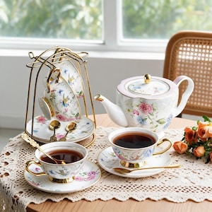 May include: A porcelain tea set featuring a teapot, cups, and saucers with a floral and hummingbird pattern, gold rims, and gold-toned spoons. The set includes a gold metal display stand holding two cups and saucers, with two additional cups and saucers.