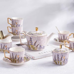 May include: A complete lavender-themed tea set with a teapot, cups, saucers, sugar bowl, and creamer. The set features a light purple background with lavender sprigs and bicycle illustrations. Gold accents adorn the handles and lids.