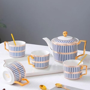 May include: A white porcelain tea set with blue and gold vertical stripes. The set includes a teapot, four teacups, and spoons. The teacups have gold handles and the teapot has a gold knob on the lid. The set is arranged on a white tray.