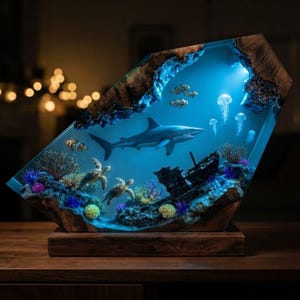 May include: A trapezoidal resin and wood lamp featuring an illuminated underwater scene with a shark, two sea turtles, clownfish, and jellyfish. The base is made of natural wood, and the interior displays a shipwreck surrounded by colorful coral.