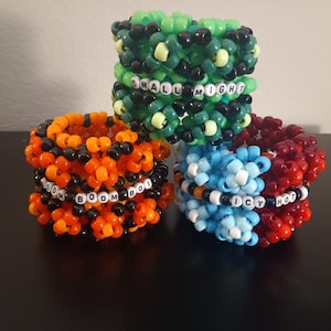 May include: Three colorful kandi bracelets stacked together. One is green with black beads and the text "SMALL MIGHT". Another is orange and black with "BOOM BOI". The last is blue and red with "ICY HOT".