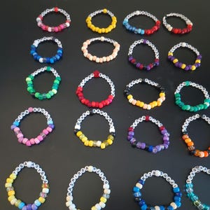 May include: A collection of colorful beaded bracelets, each with a unique design. The bracelets feature a variety of colors, including red, yellow, blue, and green, and some have white letter beads spelling out names. The bracelets are arranged on a dark surface.