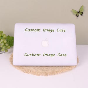 May include: A white laptop case featuring the text Custom Image Case printed in green above and below a central Apple logo. The case has a smooth finish and is designed to fit over the lid of a laptop, providing a clean and simple appearance.