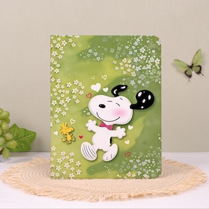 Op de afbeelding: Een rechthoekig notitieboek met een groene achtergrond en witte bloemmotieven, voorzien van de witte Snoopy-figuur met een roze vlinderdas en het kleine gele Woodstock-figuurtje, versierd met diverse kleine witte en rode hartjes.