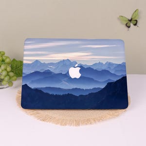 May include: A laptop skin featuring a panoramic mountain landscape with layered blue peaks and a soft, pale sky. The design displays a gradient of deep navy blue in the foreground transitioning to lighter blue ridges, centered with a white Apple logo.