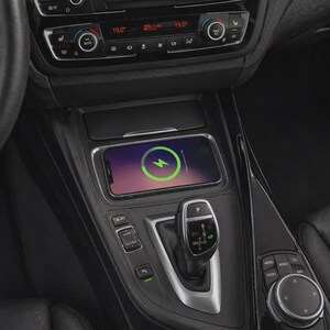 Puede incluir: Consola de coche negra con un smartphone cargando de forma inalámbrica. El teléfono muestra un símbolo de carga verde. La consola tiene botones, una palanca de cambios y una perilla de control multimedia. Se ven los ajustes del climatizador del coche.