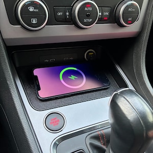 Può includere: Uno smartphone in carica wireless nella console centrale di un'auto. Lo schermo del telefono mostra un simbolo di carica. La console presenta comandi del climatizzatore e un pulsante start/stop.