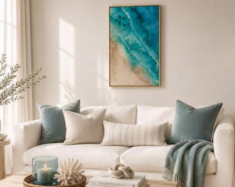 May include: A rectangular wall art print featuring an aerial view of a sandy beach meeting turquoise ocean waves. The artwork is framed in a thin gold border and hangs above a white sofa decorated with blue and beige throw pillows and a throw blanket.
