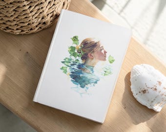 May include: A white hardcover journal featuring a watercolor illustration of a woman's profile emerging from water, surrounded by green foliage. The journal is placed on a wooden surface, next to a woven basket and a seashell.