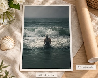 May include: A rectangular art print featuring a photograph of a person standing in the ocean waves with a dark, overcast sky. The print is displayed with a white border, accompanied by a cardboard mailing tube and a small white seashell.