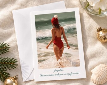 May include: A white greeting card featuring a photograph of a person in a red swimsuit and red beanie walking into ocean waves. The card includes the printed text: Jacqueline. Christmas swims with you are my favourite. A white envelope is included.