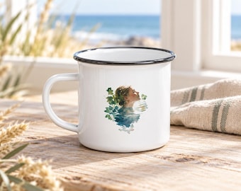 May include: A white enamel mug with a black rim and a handle on the left side. The mug features a watercolor-style illustration of a person partially submerged in water, surrounded by green foliage, positioned on the center of the mug's exterior.