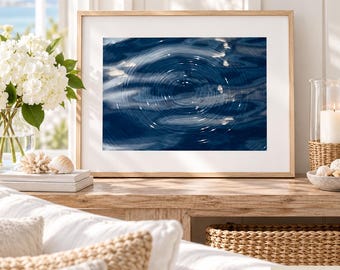 May include: A framed horizontal wall art print featuring a deep blue water surface with concentric ripples and light reflections. The print is displayed in a light-colored wooden frame with a white mat, positioned on a wooden surface.