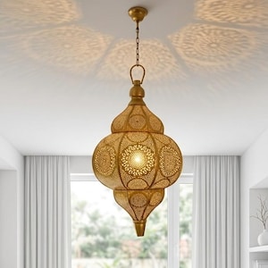 May include: A gold-colored, ornate pendant light fixture with a Moroccan design. The lamp is suspended from the ceiling by a chain and features intricate cut-out patterns that cast decorative light patterns. The fixture has a bulb inside, and is in a room with white curtains.