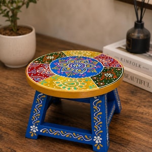 May include: A small wooden stool with a round top featuring a colorful hand-painted mandala design in blue, yellow, red, and green. The stool has four sturdy blue legs decorated with white floral patterns and yellow accents along the edges.