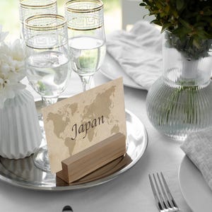 May include: A table setting with four clear wine glasses with gold rims, a place card that says "Japan", and a vase of white flowers. The setting is on a silver tray with a white tablecloth.