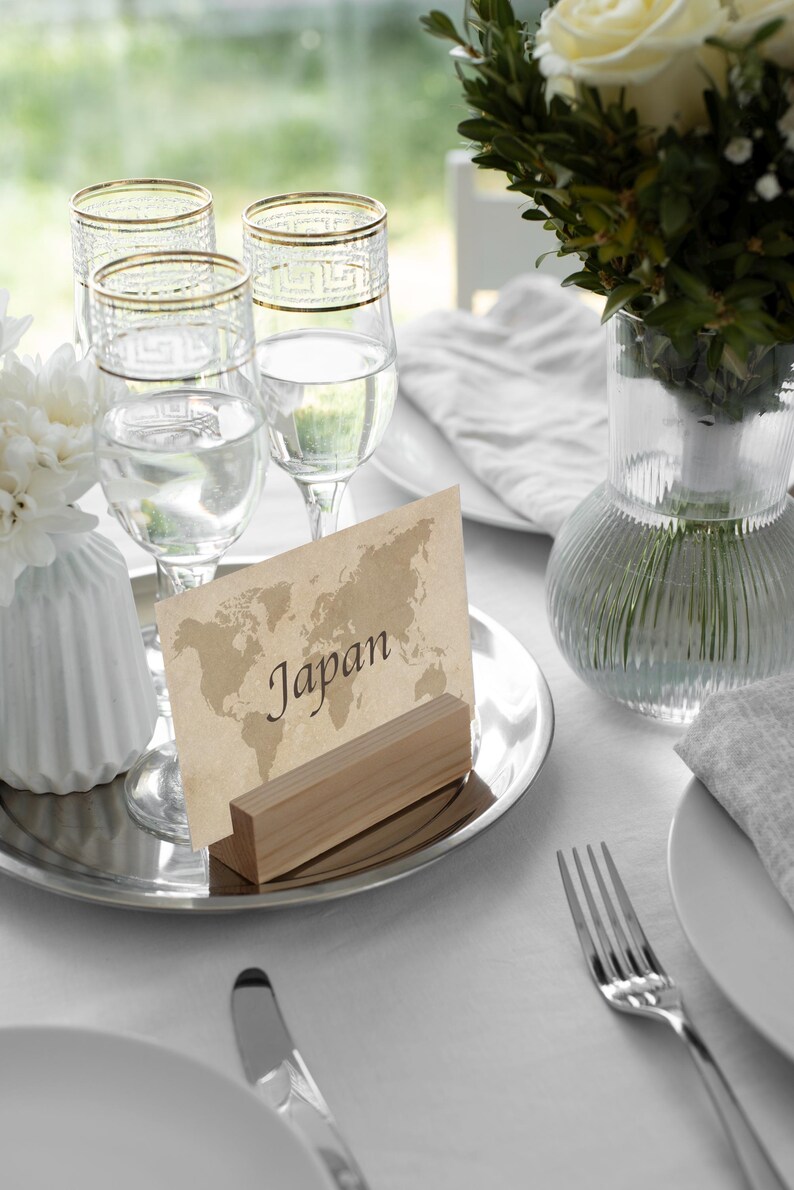 May include: A table setting with four clear wine glasses with gold rims, a place card that says "Japan", and a vase of white flowers. The setting is on a silver tray with a white tablecloth.