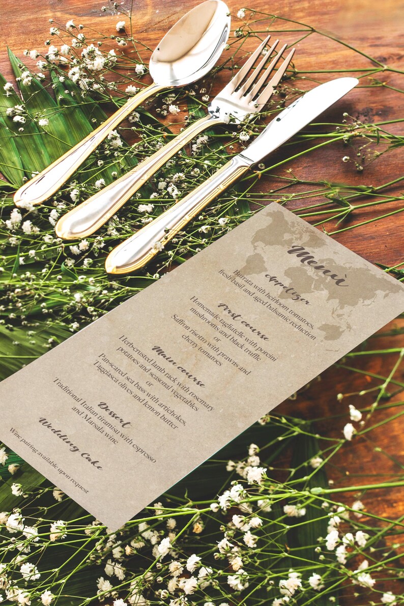 May include: A formal table setting with silverware, including a spoon, fork, and knife, with gold accents. The utensils are arranged on a wooden surface, decorated with greenery and a menu card.