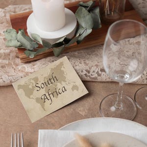 May include: A table setting with a lit white candle, greenery, and a card that reads "South Africa" with a world map. A wine glass, plate, and silverware are also present, creating an elegant ambiance.