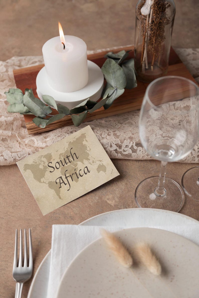 May include: A table setting with a lit white candle, greenery, and a card that reads "South Africa" with a world map. A wine glass, plate, and silverware are also present, creating an elegant ambiance.