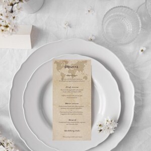 May include: A wedding menu card on a white plate, with a floral arrangement and a candle. The menu card features a world map design and a list of courses. The overall theme is elegant and romantic.