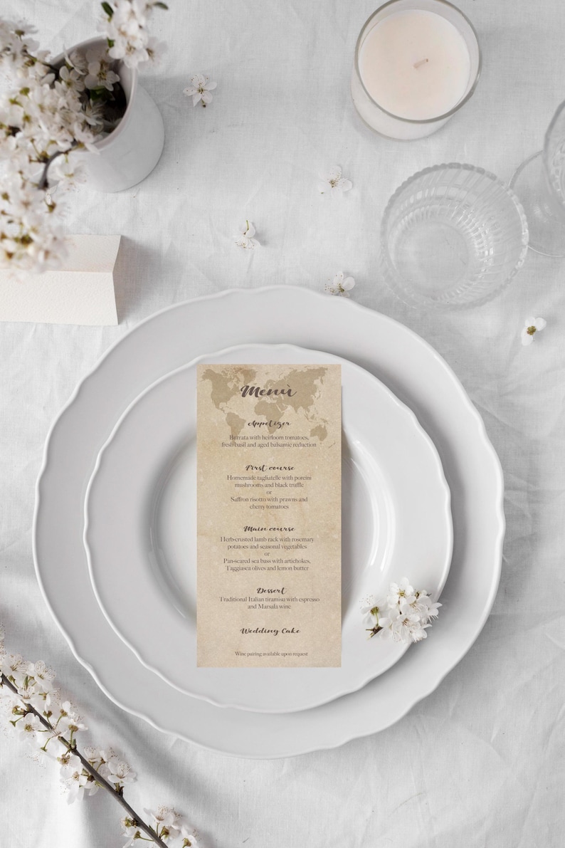 May include: A wedding menu card on a white plate, with a floral arrangement and a candle. The menu card features a world map design and a list of courses. The overall theme is elegant and romantic.