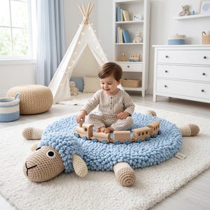 Pode incluir: Um tapete de ovelha de crochê azul claro com rosto e pernas bege. Um comboio de brinquedo de madeira está no tapete. O tapete está sobre um tapete branco e fofo no quarto de uma criança. Uma criança brinca com o comboio.