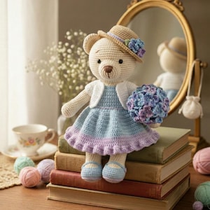 May include: A handmade crochet bear doll wearing a blue and purple dress, a white cardigan, and a straw hat with a flower. The bear is holding a bouquet of blue and purple flowers and is sitting on a stack of books. The text "Handmade Crochet Bear" is at the bottom.