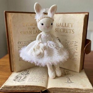Puede incluir: Una muñeca de conejo de ganchillo blanco hecha a mano que lleva un vestido y sostiene un bolso, con el texto "Bunny Crochet Pattern" arriba. El conejo tiene orejas largas y posa frente a un libro abierto con las palabras "Stories from Swan Lake" y "Ballet Secrets".