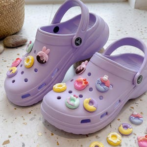 May include: A pair of light purple slip-on clogs featuring a series of circular ventilation holes on the upper. Each shoe is decorated with various colorful donut-shaped charms, including designs with small animal ears and pastel icing details.