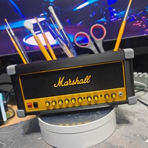 Op de afbeelding: Zwarte en gele bureau-organizer in de vorm van een Marshall-versterker, met het woord "Marshall" in gele letters. De organizer bevat penselen, pennen en een schaar. De voorkant heeft knoppen en een aan/uit-knop.