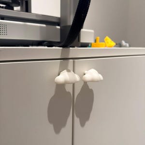 Pode incluir: Dois puxadores de armário brancos em forma de nuvem presos a um armário cinza claro. Os puxadores projetam sombras em forma de nuvem nas portas do armário. Uma impressora 3D é visível ao fundo.