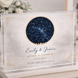Puede incluir: Un bloque acrílico transparente con un diseño de mapa celeste. Presenta un círculo azul oscuro con un patrón de constelaciones, detalles dorados y los nombres "Emily & James" con una fecha y coordenadas. También incluye el texto "On the night we became us".