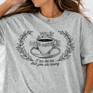Op de afbeelding: Een heather grijze t-shirt met een zwart-witte illustratie van een theekop en schotel, omringd door een bloemenkrans. De tekst "I am the tea that you are having" staat eronder.