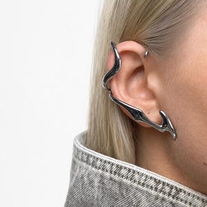 May include: A silver-toned ear cuff with a fluid, organic, and elongated shape that wraps around the outer edge of the ear. The metal surface is smooth and reflective, featuring a continuous, undulating design that extends from the upper cartilage down to the earlobe.