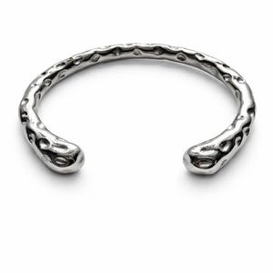 Puede incluir: Un brazalete abierto de color plateado con una superficie texturizada y martillada. El brazalete tiene una forma redondeada con extremos lisos y bulbosos. El metal tiene un aspecto ligeramente irregular y orgánico.