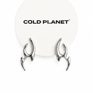 May include: A pair of silver-toned earrings with a curved, flame-like design attached to a circular white display card featuring the black text COLD PLANET. The earrings have a polished, metallic finish and a sharp, abstract shape.
