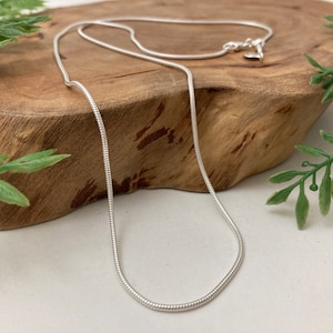 May include: A silver snake chain necklace on a wooden surface. The chain is thin and delicate, and it is perfect for layering with other necklaces.